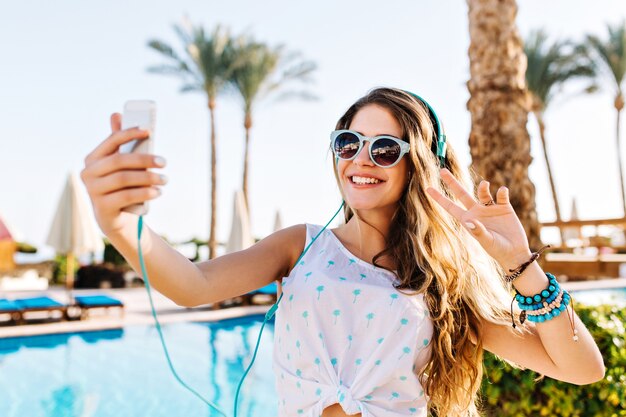 Happy girl in sunglasses with tanned skin making selfie with peace sign on palm trees background