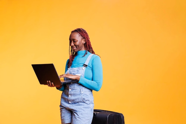 Free photo happy girl using laptop on camera over orange background, browsing online website in studio. female traveller with luggage holding wireless pc to check social media internet page.