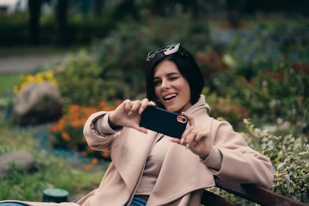 Счастливая девушка с помощью смартфона в городском парке, сидя на скамейке