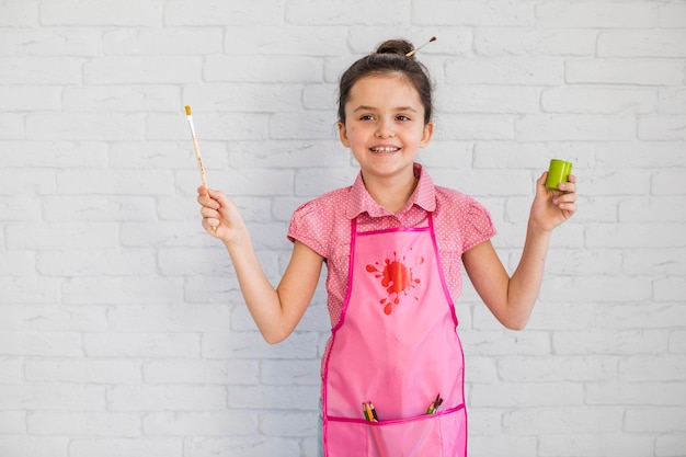 Ragazza felice che indossa il pennello rosa della tenuta del grembiule e la bottiglia della pittura a disposizione