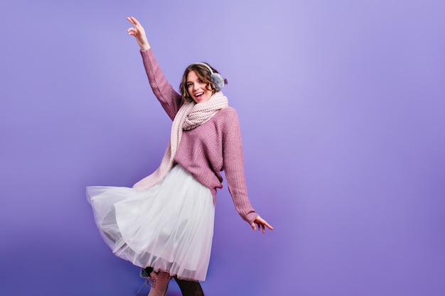 Happy girl in white knitted scarf and fur headphones dancing and waving hands. Indoor photo of fashionable caucasian woman wears scarf and winter accessories isolated on purple wall with laugh.