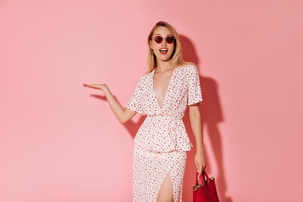 Happy girl with blonde hair in red glasses and white dress holding cool handbag and pointing to place for text on pink background