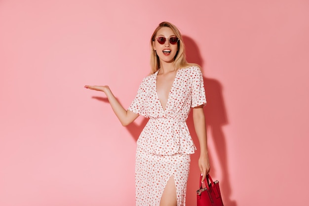 Happy girl with blonde hair in red glasses and white dress holding cool handbag and pointing to place for text on pink background