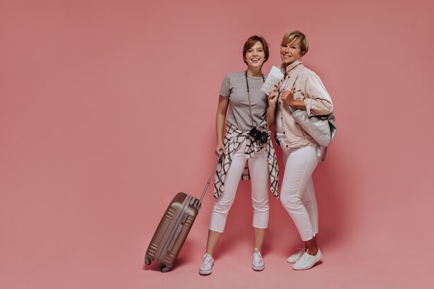 Happy girl with dark hair in light trousers and grey t-shirt holding suitcase, tickets and camera and posing with smiling woman on pink backdrop. 