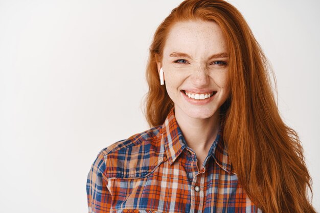 Happy girl with ginger hair and freckles smiling enjoy listening music in wireless earphones having fun standing over white background