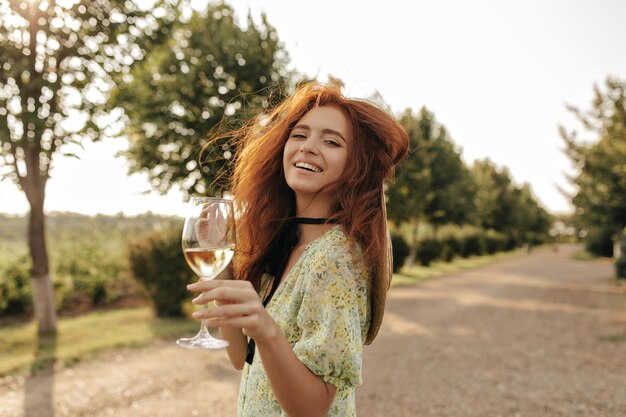Happy girl with long ginger hair in summer yellow outfit smiling looking into camera and holding glass with champagne outdoor