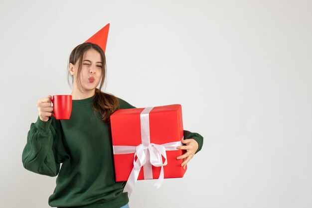 happy girl with party cap holding her christmas gift and a cup on white