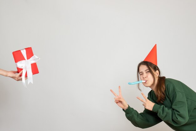 happy girl with party cap making victory sign using noisemaker human hand holding gift on white