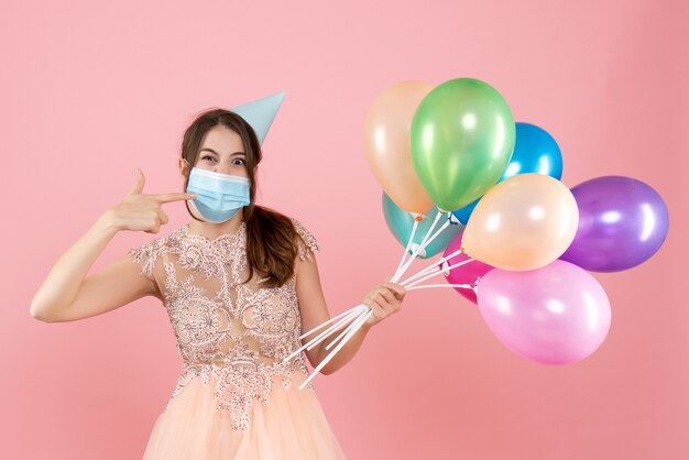 ピンクのカラフルな風船を保持している彼女のマスクを指しているパーティーキャップと医療マスクを持つ幸せな女の子