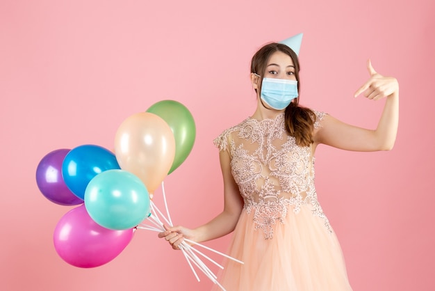 ピンクのカラフルな風船を持って自分を指しているパーティーキャップと医療マスクを持つ幸せな女の子