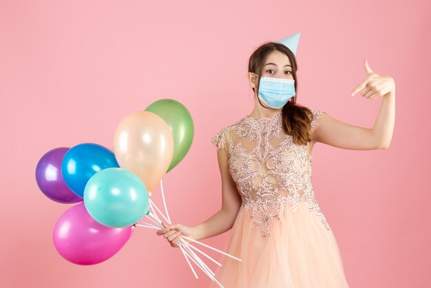 happy girl with party cap and medical mask pointing at herself holding colorful balloons on pink