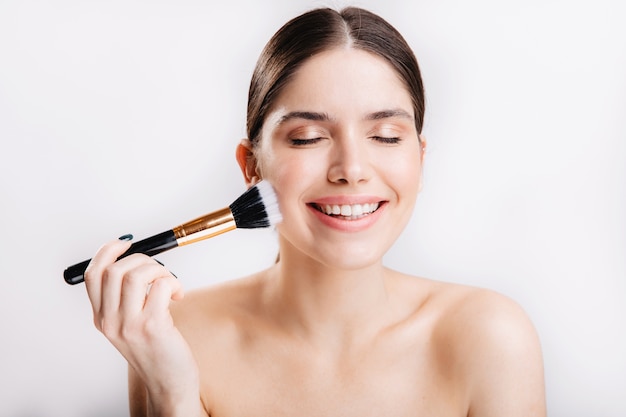 Happy girl with perfect healthy skin is smiling and touching her face with brush on white wall.