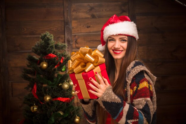 Счастливая девушка с красными губами держит подарок возле новогодней и рождественской елки. Красивая дама в шляпе Санты смотрит в камеру.