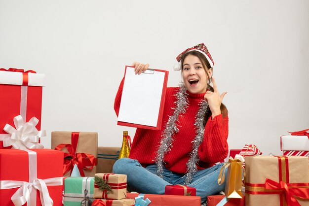 happy girl with santa hat holding files sitting around presents on white