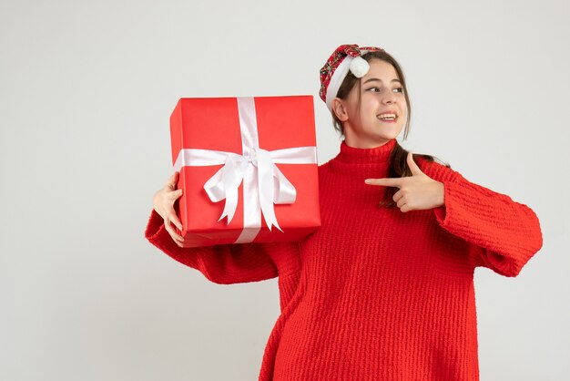 happy girl with santa hat holding present finger pointing the box standing on white