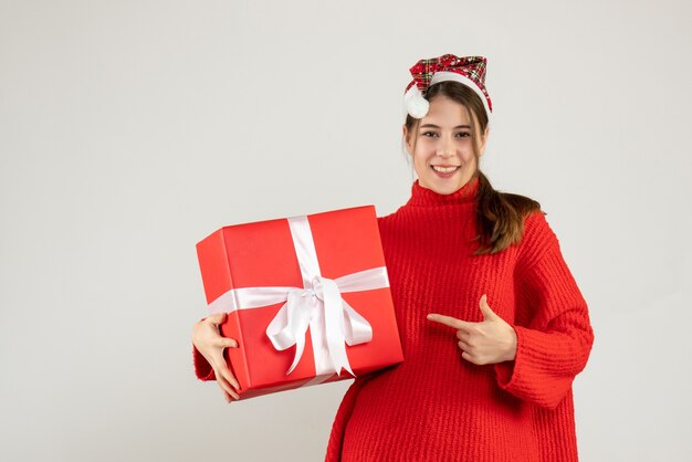 happy girl with santa hat holding present finger pointing her gift standing on white