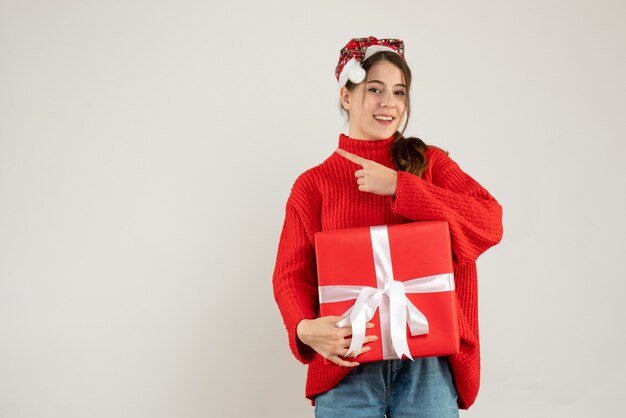 happy girl with santa hat holding present finger pointing something standing on white