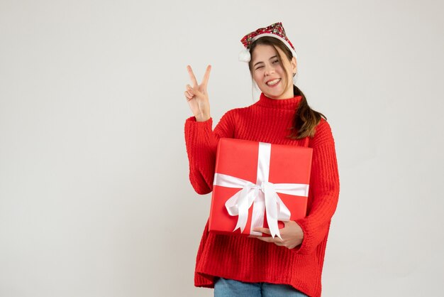 happy girl with santa hat holding present making victory sign standing on white