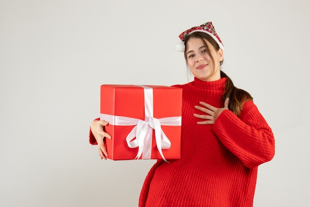 happy girl with santa hat holding present putting hand on her chest standing on white