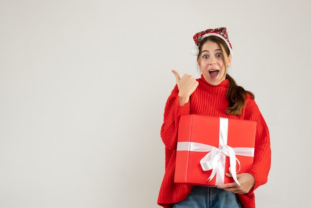 happy girl with santa hat holding present showing something standing on white