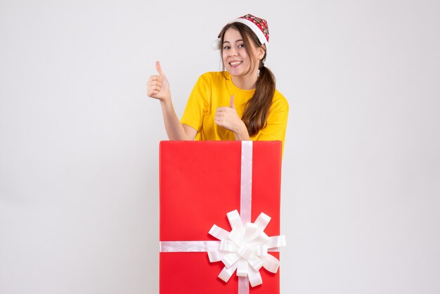 白の大きなクリスマスプレゼントの後ろに立っているサインアップサインを作るサンタ帽子と幸せな女の子