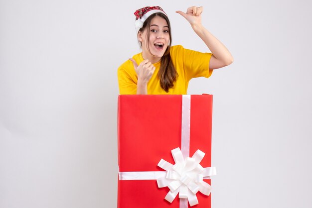 happy girl with santa hat making thumb up sign standing behind big xmas gift on white