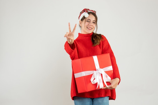 happy girl with santa hat making victory sign holding gift on white