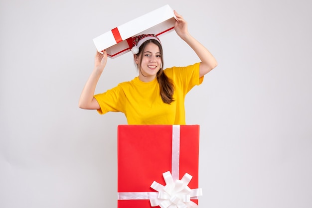 Ragazza felice con il cappello della santa che alza la copertura della scatola sopra la sua testa che sta dietro il grande regalo di natale su bianco