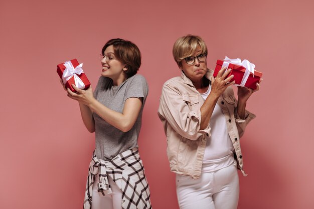 Happy girl with short hair holding gift box, rejoicing and posing with stylish surprised old lady in white outfit on isolated backdrop. 