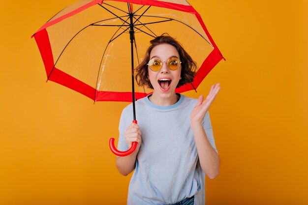 Happy girl with short hairstyle posing with laugh under umbrella. Studio shot of enchanting white woman in trendy sweater holding parasol on yellow wall.
