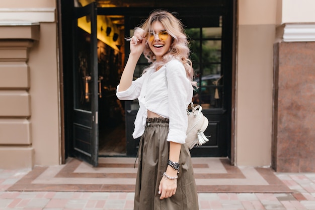 Foto gratuita ragazza felice con l'acconciatura riccia alla moda che ride e che tiene gli occhiali da sole gialli mentre posa davanti al negozio