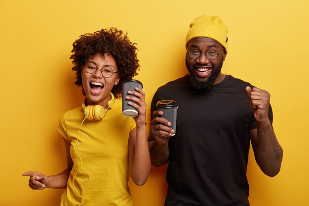 Happy girlfriend and boyfriend move actively, dance and have fun drink coffee to go, wear casual clothes, use stereo headphones, isolated over yellow wall. People, leisure and lifestyle concept