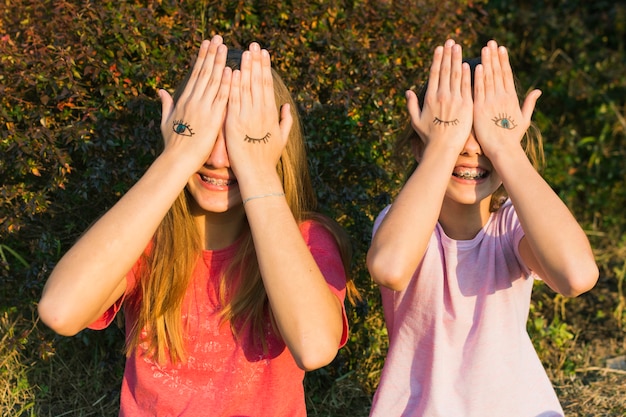 Happy girls standing in front of plant covering their eyes with tattoo on hand
