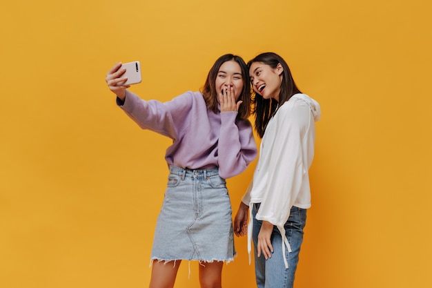 Happy girls in sweatshirts take selfie and laugh on orange wall