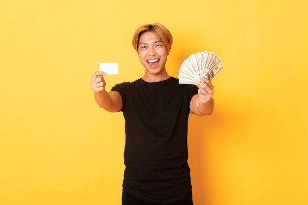 お金とクレジットカードを示す黒のカジュアルな服装で幸せそうなアジアの男、生意気な黄色の壁を笑顔