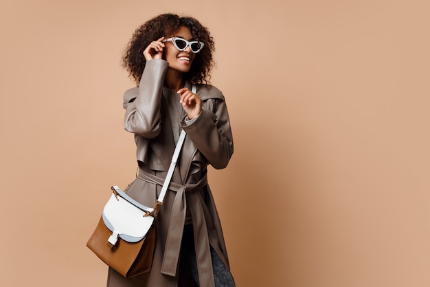 Happy good looking black woman wearing grey leather coat , posing on beige background. Autumn or winter fashion concept.