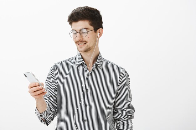 happy good-looking caucasian bearded guy in round glasses, holding smartphone and listening music in earphones, using gadget to find cafe on map, standing carefree and relaxed