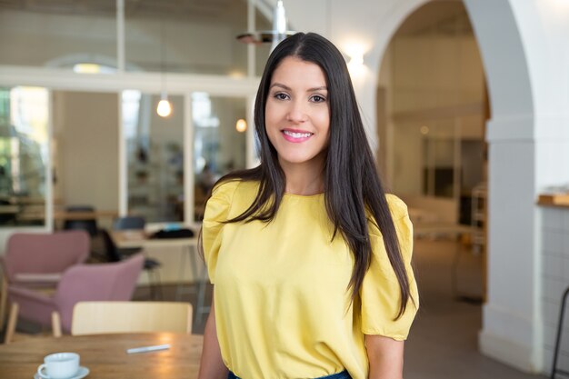 Happy gorgeous black haired woman wearing yellow shirt, standing in co-working space, posing, 