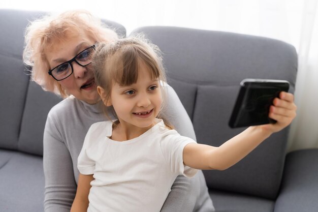 Счастливая бабушка и внучка вместе пользуются телефоном, сидят дома на уютном диване, просматривают приложения для мобильных устройств, бабушка с внуком играют в игры, смотрят на экран, веселятся.