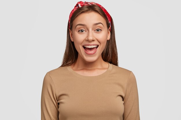 Happy green eyed European woman with pleased expression, keeps mouth opened, wears red bandana