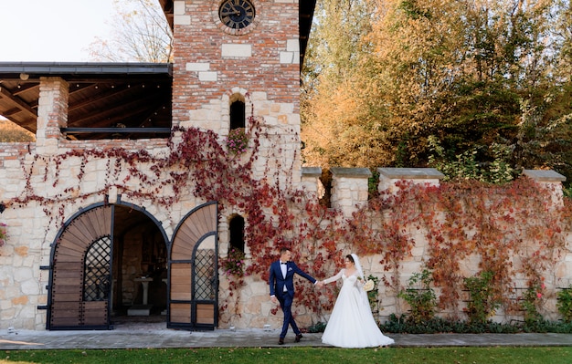 Happy groom and bride are holding hands together and walking in front of old stone building on the warm autumn day