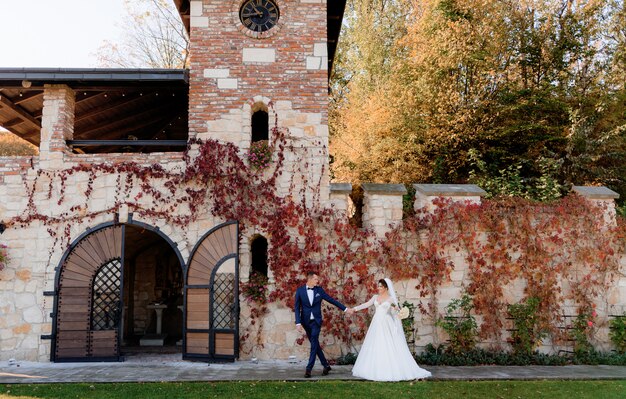 Счастливые жених и невеста вместе держатся за руки и гуляют перед старым каменным зданием в теплый осенний день