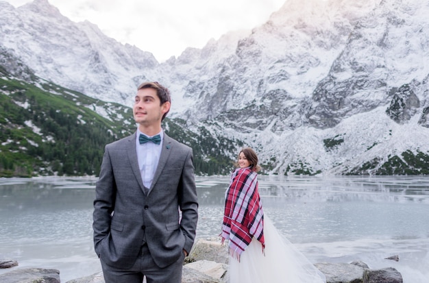 Foto gratuita lo sposo e la sposa felici si distinguono vicino al lago ghiacciato circondato da montagne innevate