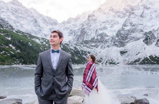 행복한 신랑과 신부는 눈 덮인 산으로 둘러싸인 얼어 붙은 호수 근처에 떨어져 서있다