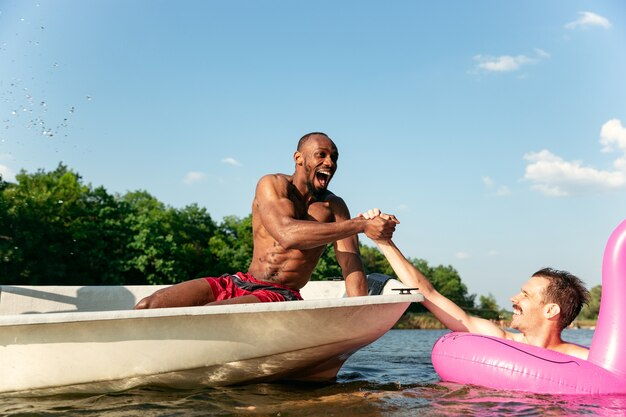 笑ったり、水をはねたり、川で泳いだりしながら楽しんでいる友達の幸せなグループ。晴れた日に川沿いでボートに乗って水着姿のうれしそうな男性。夏、友情、リゾート、週末のコンセプト。