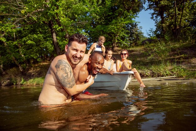 笑ったり川で泳いだりしながら楽しんでいる友達の幸せなグループ。晴れた日に川沿いのボートで水着姿のうれしそうな男女。夏、友情、リゾート、週末のコンセプト。
