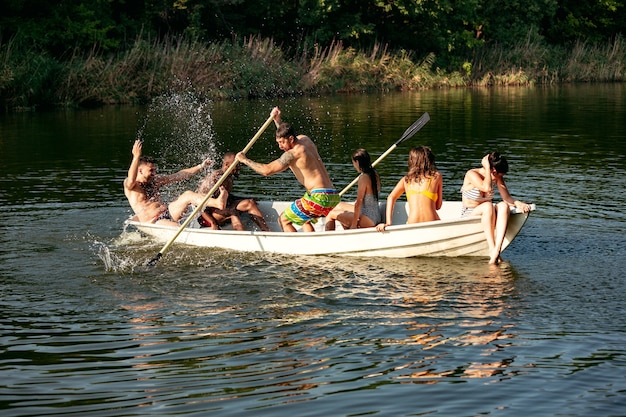 Счастливая группа друзей весело смеясь и купаясь в реке. Радостные мужчины и женщины в купальнике в лодке на берегу реки в солнечный день. Лето, дружба, курорт, концепция выходных.