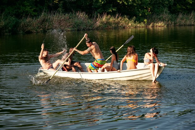 Happy group of friends having fun while laughting and swimming in river. Joyful men and women in swimsuit in a boat at riverside in sunny day. Summertime, friendship, resort, weekend concept.