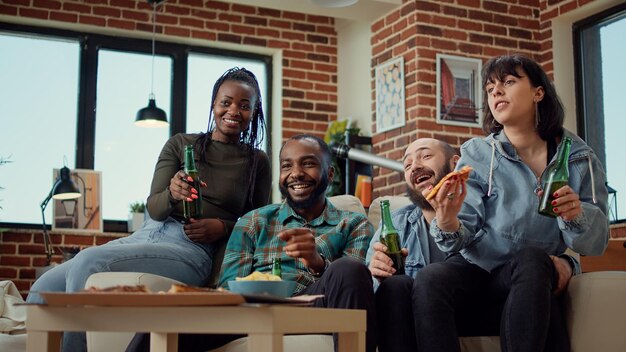 Happy group of friends making toast with beer bottles and watching comedy movie on television, leisure activity. Diverse people having fun with alcohol and film on tv channel at home.