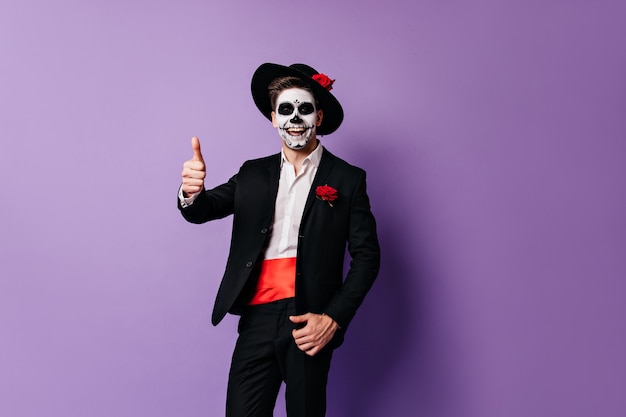 Happy guy in mexican clothes and mask laughs and shows thumb up on purple background.